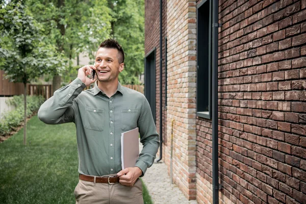 Pleased real estate agent with folder looking away while talking on mobile phone near cottage — Stock Photo