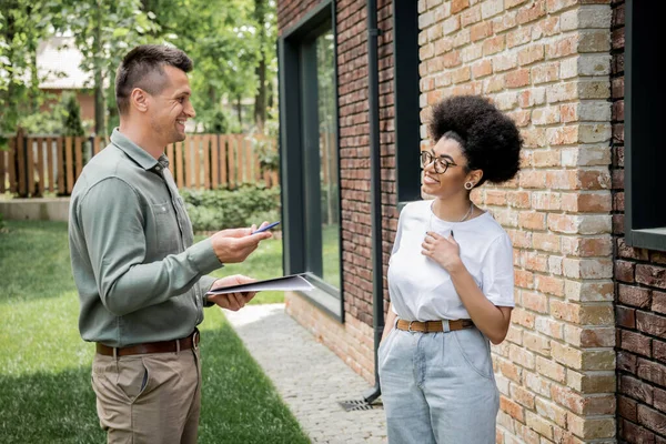 Agente imobiliário com pasta e caneta conversando com o cliente afro-americano sorridente perto de nova casa de campo — Fotografia de Stock