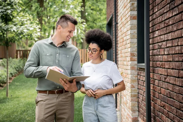 Smiling real estate agent showing documents to african american woman near new city cottage — Stock Photo
