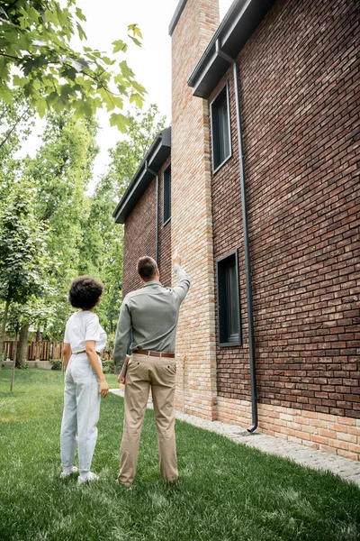 Vue arrière du courtier immobilier pointant avec la main et montrant une maison neuve à un client afro-américain — Photo de stock