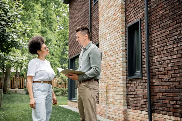 Agent immobilier confiant avec dossier parlant à l'acheteur afro-américain positif près de nouveau chalet — Photo de stock