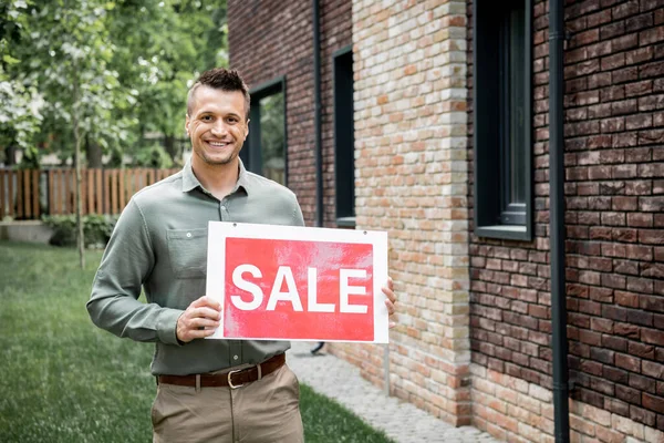 Agente de bienes raíces feliz y seguro de sí mismo mirando a la cámara y sosteniendo letrero de venta cerca de casa nueva - foto de stock