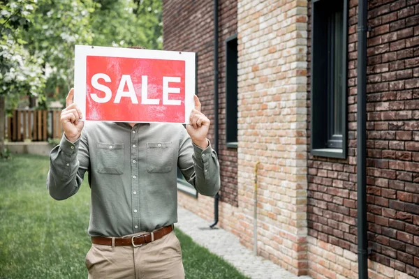 Агент по недвижимости скрывает лицо вывеской для продажи, стоя рядом с коттеджем в городе — стоковое фото