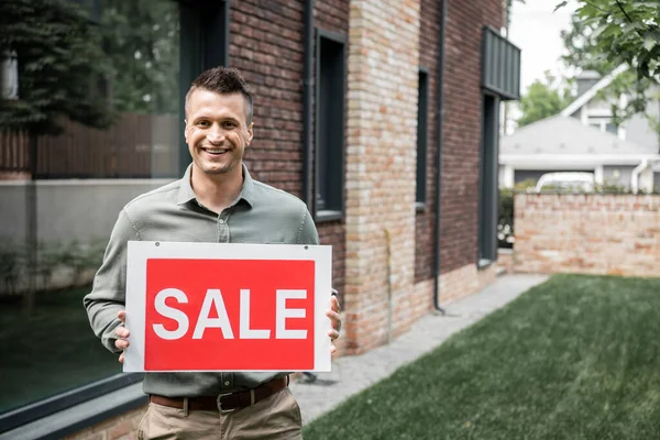 Optimistic realtor holding signboard with sale lettering and looking at camera near cottage — Stock Photo
