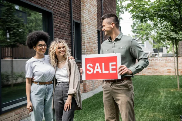 Real estate agent holding sale signboard near cheerful interracial lesbian women and new cottage — Stock Photo