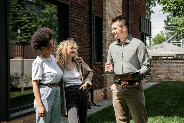 Agente imobiliário com pasta conversando com casal interracial lésbico sorridente perto de edifício na rua — Fotografia de Stock