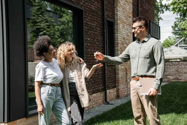 Corretor imobiliário com pasta dando chave de casa nova para casal lésbico multiétnico feliz — Fotografia de Stock