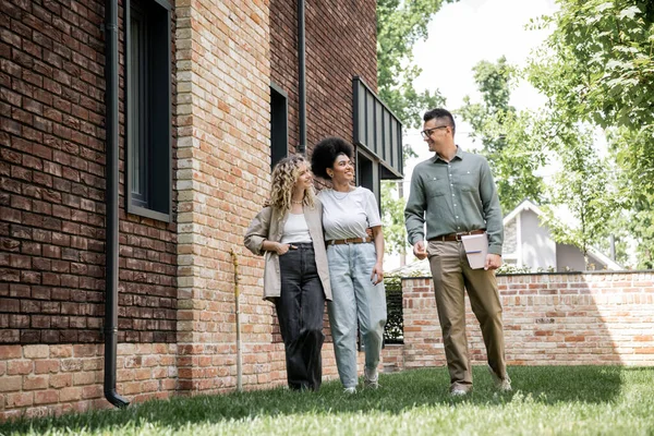 Full length of real estate agent and multiethnic lesbian couple walking and talking near new cottage — Stock Photo