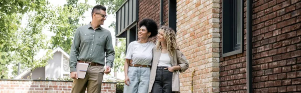 Joyful multicultural lesbian couple talking with real estate agent near house on street, banner — Stock Photo