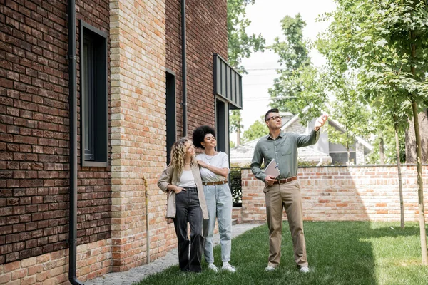 Agente imobiliário com pasta apontando com a mão perto de casa de campo e casal lésbico interracial — Fotografia de Stock