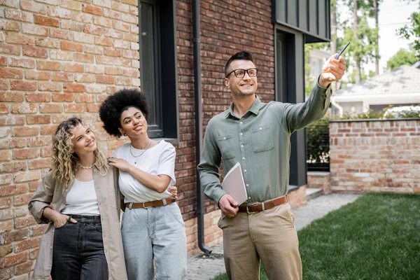 Cheerful interracial lesbian women looking at realtor pointing with pen near house on street — Stock Photo