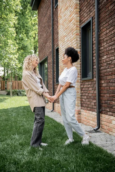 Side view of delighted multiethnic lesbian couple holding hands near new private cottage — Stock Photo