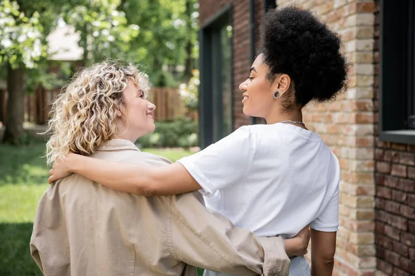 Cheerful lesbian couple embracing and smiling at each other next to new own house outdoors — Stock Photo
