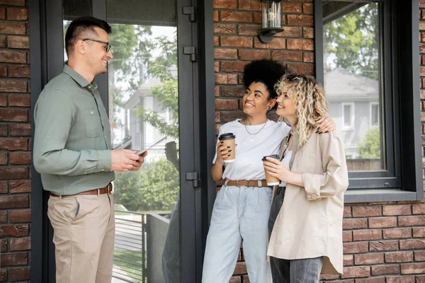 Real estate broker talking to happy interracial lesbian couple with paper cups near new cottage — Stock Photo