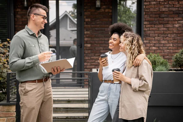 Cheerful multiethnic lesbian couple with coffee to go talking with real estate agent near new house — Stock Photo