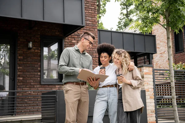 Positive realtor showing documents to interracial lesbian couple with paper cups near house — Stock Photo