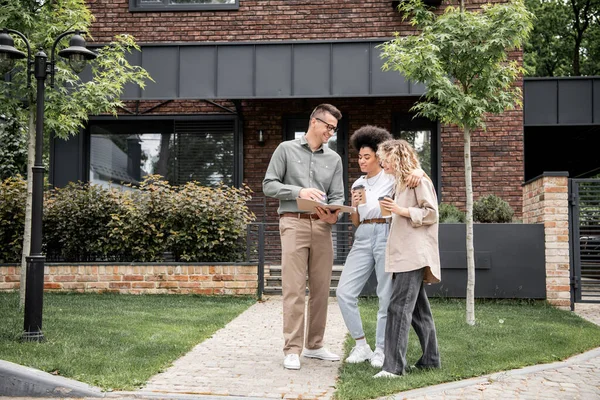 Confident real estate broker showing documents to multiethnic lesbian couple near modern cottage — Stock Photo