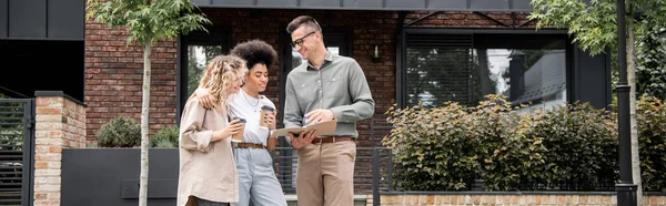 Sonriente agente inmobiliario mostrando documentos a interracial pareja del mismo sexo con vasos de papel al aire libre, pancarta - foto de stock