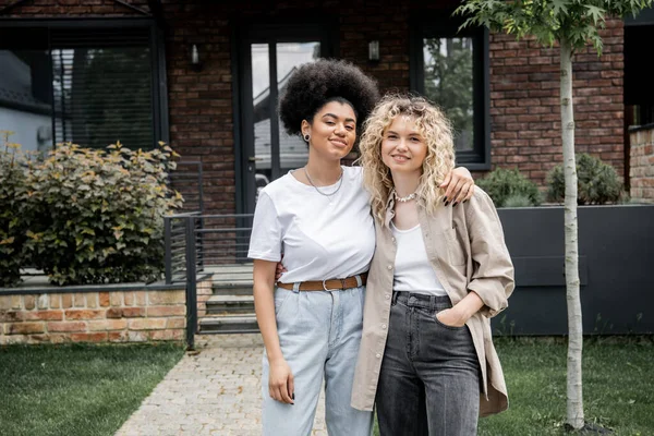 Pleased multiethnic lesbian couple embracing and looking at camera near new private house — Stock Photo