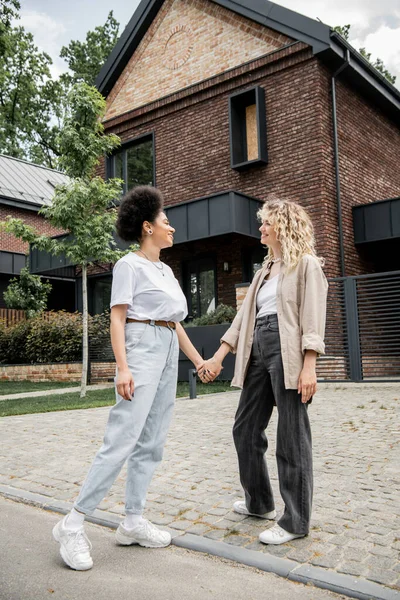 Longitud completa de la pareja lesbiana multicultural tomados de la mano y sonriendo el uno al otro cerca de su propia casa de campo - foto de stock