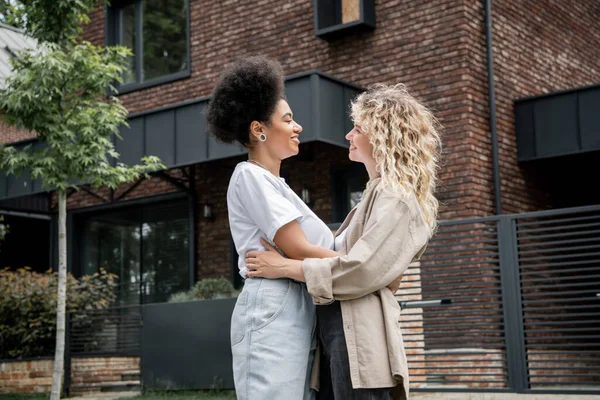 Side view of interracial lesbian couple embracing and smiling at each other near new own cottage — Stock Photo