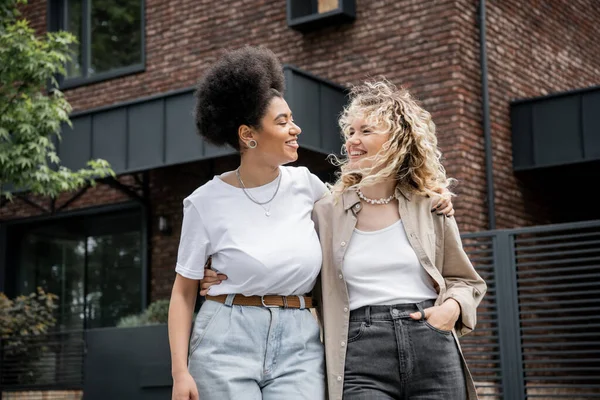Overjoyed multiethnic lesbian couple hugging and looking at each other next to private house — Stock Photo
