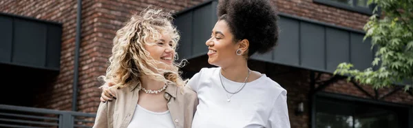 Happy and successful interracial lesbian couple looking at each other near own cottage, banner — Stock Photo
