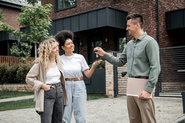 Exitoso agente inmobiliario con carpeta que da la llave de la nueva casa a la pareja lesbiana multiétnica complacida - foto de stock