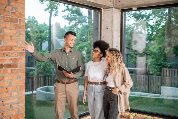 Sonriente agente inmobiliario apuntando con la mano y mostrando el interior de un nuevo hogar a una pareja lesbiana interracial - foto de stock