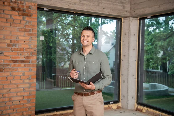 Happy successful real estate broker holding folder and pen while standing inside new cottage — Stock Photo