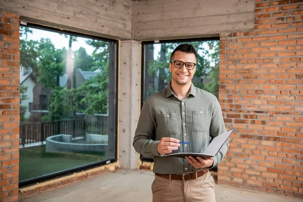 Joyful real estate agent in eyeglasses holding folder with pen and looking at camera in new house — Stock Photo