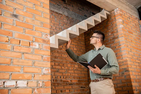 Immobilienmakler mit Brille hält Ordner in der Hand und berührt Treppen im Haus mit unfertigem Interieur — Stockfoto