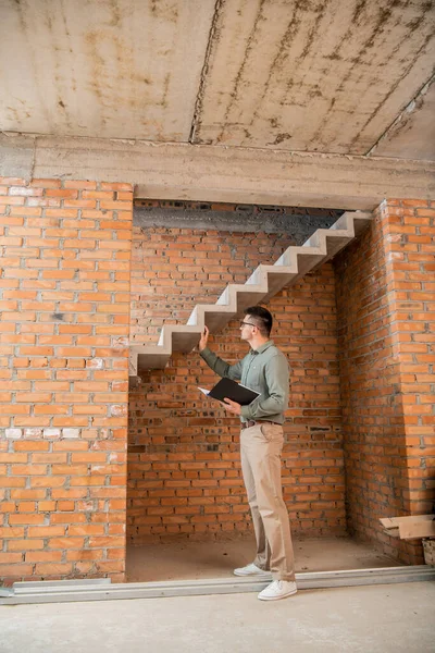 Pleine longueur de l'agent immobilier avec dossier touchant escaliers dans la maison avec intérieur inachevé — Photo de stock