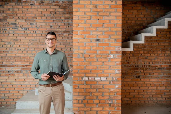 Joyful real estate agent in eyeglasses looking at camera in house with unfinished interior — Stock Photo