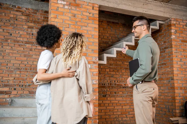 Agent immobilier pointant vers les escaliers et montrant la maison avec intérieur inachevé pour lesbienne couple interracial — Photo de stock