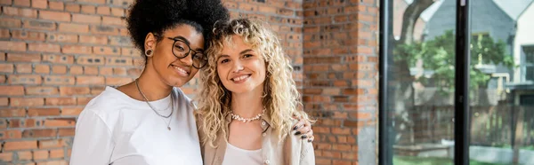 Pleased interracial lesbian couple embracing and smiling at camera in new house, banner — Stock Photo