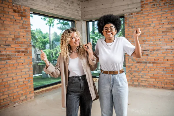 Overjoyed interracial lesbian couple holding hands and looking at camera in new private cottage — Stock Photo