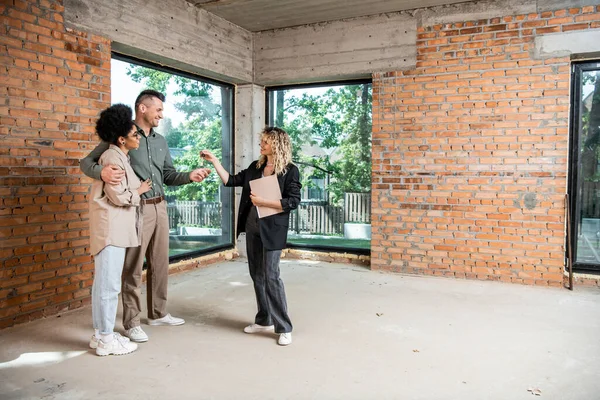 Immobilienmakler übergibt Schlüssel an gemischtrassiges Paar in neuem Ferienhaus mit unvollendetem Interieur — Stockfoto