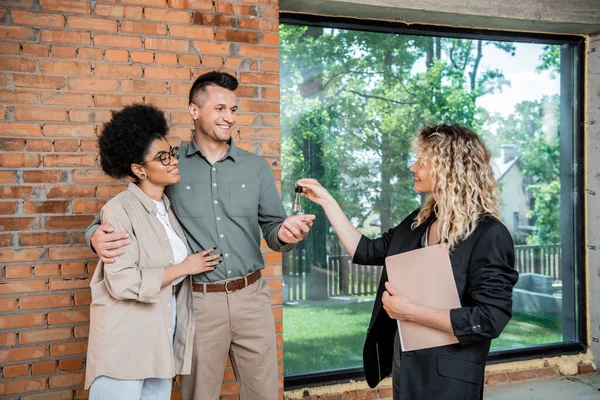 Corretor imobiliário com documentos dando chaves de casa nova para casal multicultural satisfeito — Fotografia de Stock