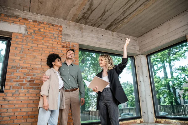 Immobilienmakler zeigt mit der Hand an die Decke, während er dem multiethnischen Paar ein neues Haus zeigt — Stockfoto