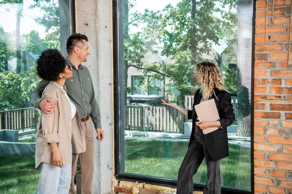 Immobilienmakler mit Ordner zeigt mit der Hand auf Fenster in neuem Ferienhaus in der Nähe gemischtrassiges Paar — Stockfoto