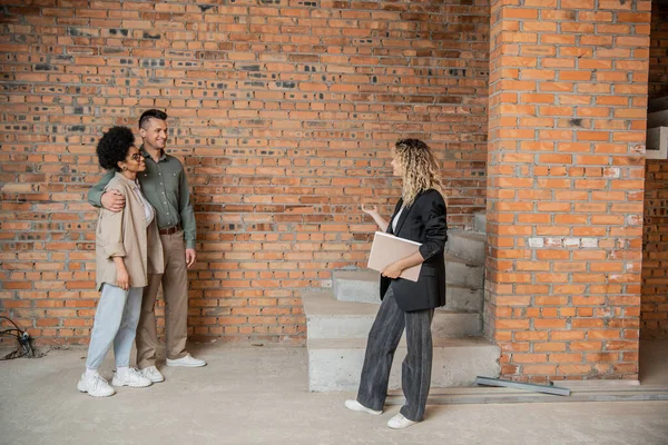 Real estate broker with folder showing unfinished interior of new house to interracial couple — Stock Photo