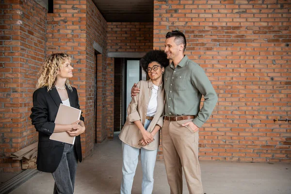 Pleased multiethnic couple embracing near real estate agent standing with folder in new cottage — Stock Photo