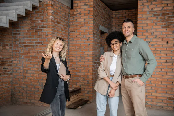 Sonriente corredor de bienes raíces señalando con la mano y mostrando nueva vivienda a la pareja multicultural - foto de stock