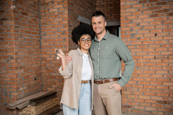 Overjoyed interracial couple with keys looking at camera in new private cottage — Stock Photo