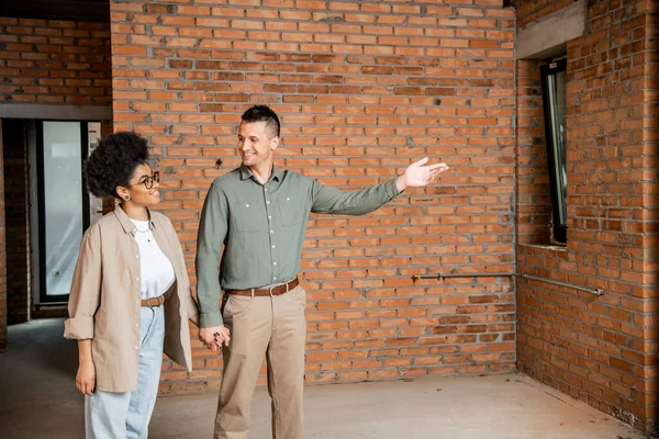 Happy man pointing with hand while showing new house to african american woman — Stock Photo