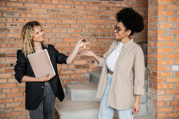 Agente de bienes raíces rubia positiva con la carpeta que da llaves de nueva casa de campo para el comprador afroamericano - foto de stock