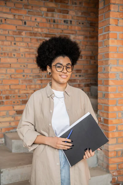 African american real estate agent in eyeglasses holding folder and smiling at camera in new house — Stock Photo