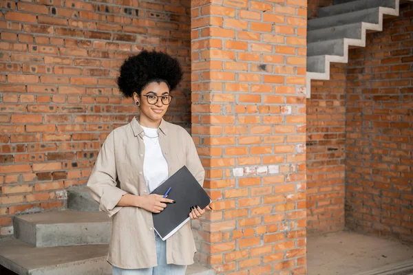 Happy african american real estate agent with folder looking at camera in new house — Stock Photo
