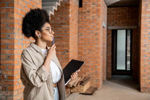 Vue de côté de réfléchi agent immobilier afro-américain debout avec stylo et dossier dans une maison neuve — Photo de stock